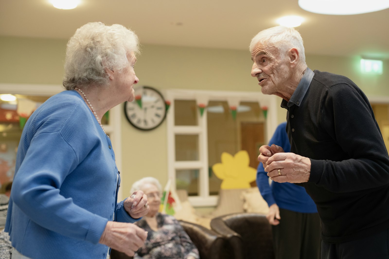 Two residents standing and talking in the common room of a senior living community.